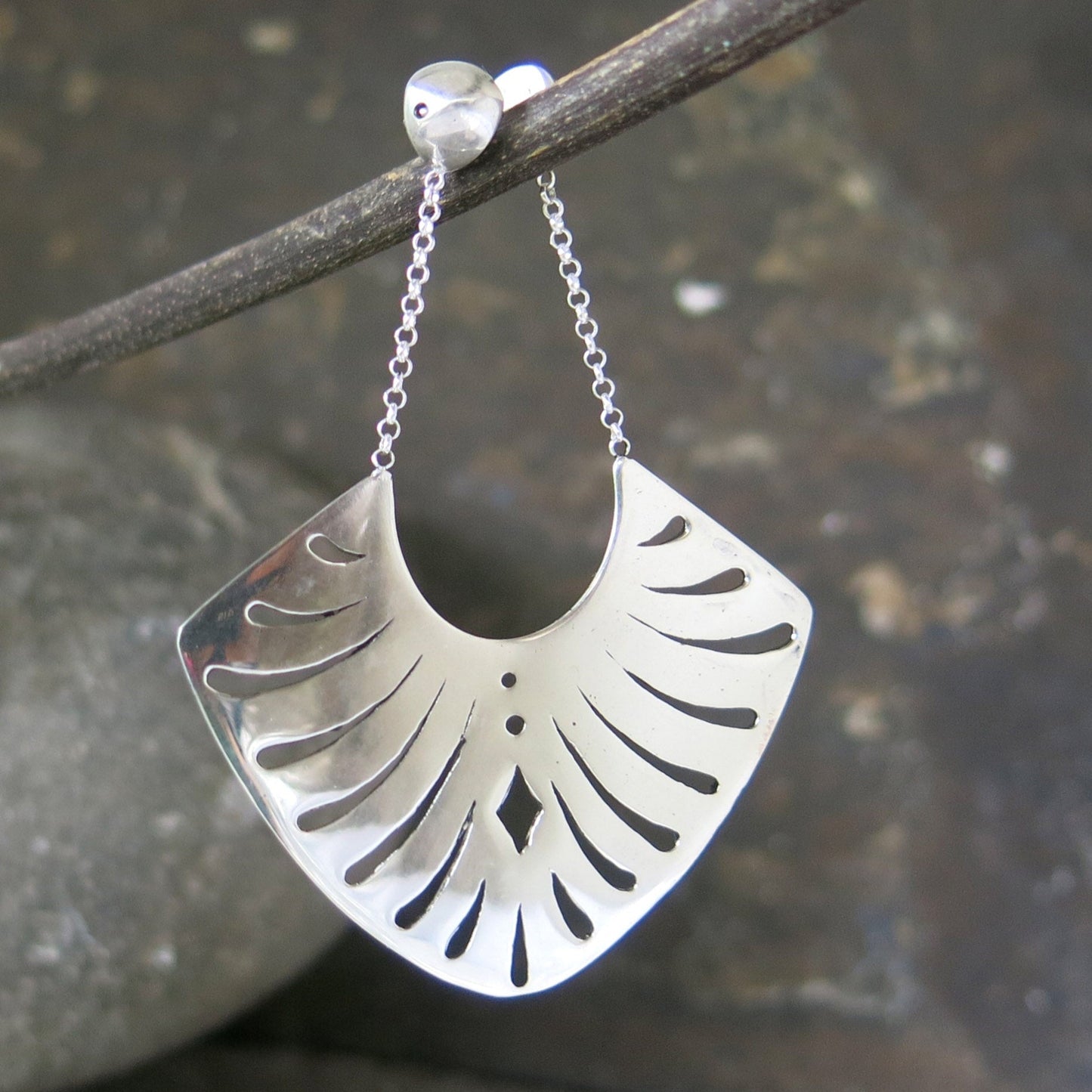 A pair of sterling silver earrings with a fan-shaped design, showcasing intricate geometric cut-out patterns. The earrings dangle from delicate silver chains attached to polished stud tops, creating a refined and sophisticated appearance. Photographed against a rustic stone backdrop to highlight their shimmering finish.