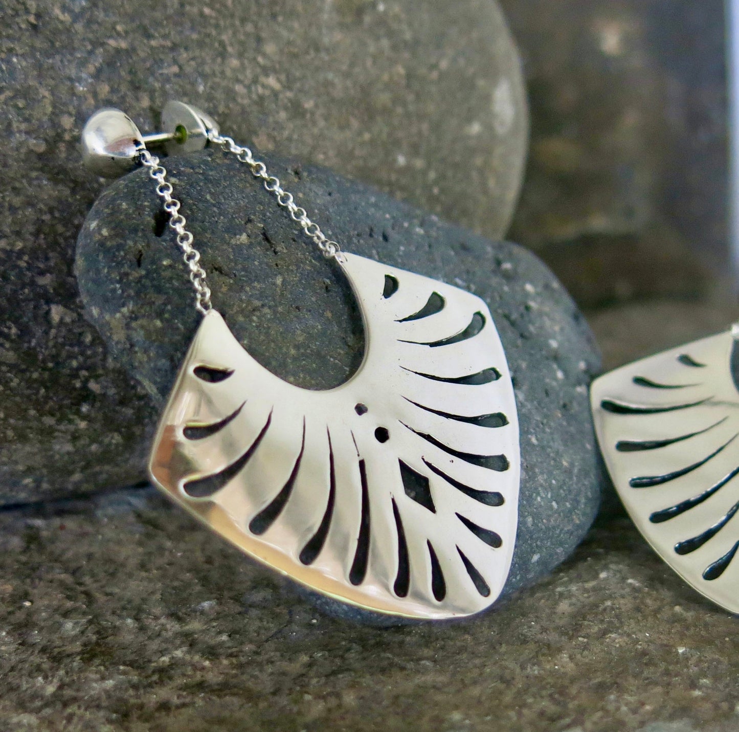 A pair of sterling silver earrings with a fan-shaped design, showcasing intricate geometric cut-out patterns. The earrings dangle from delicate silver chains attached to polished stud tops, creating a refined and sophisticated appearance. Photographed against a rustic stone backdrop to highlight their shimmering finish.